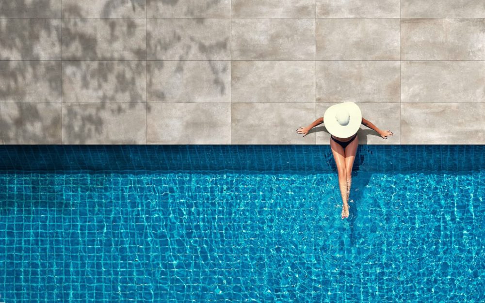 SIKO Žena v klobúku sedí na okraji bazéna s elegantnou dlažbou k bazénu v dizajne kameňa.